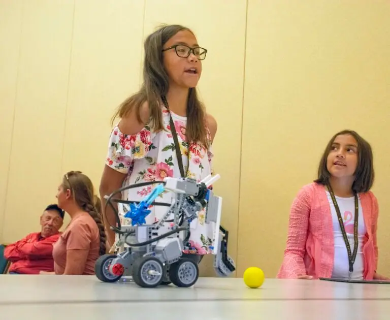 two girls stand behind their robot on a table during contest where the robot, made of legos is moving a ball