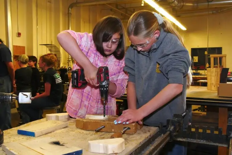 a girl drills a hole through some wood on a work bench while another girl helps keep the wood steady