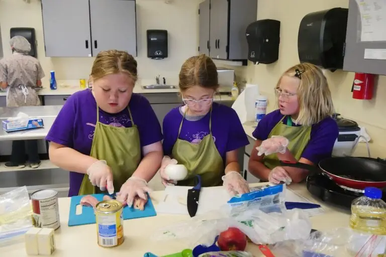 three girls stand at a counter top cutting chicken and preparing ingredients for a meal