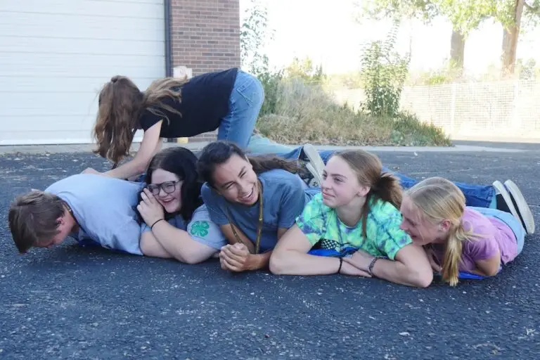 a group of smiling young people lay side by side on the ground while another climbs over the top in the middle of a game