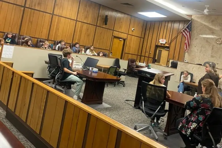 a group of young people sit in a county commission chamber listening to an adult talk