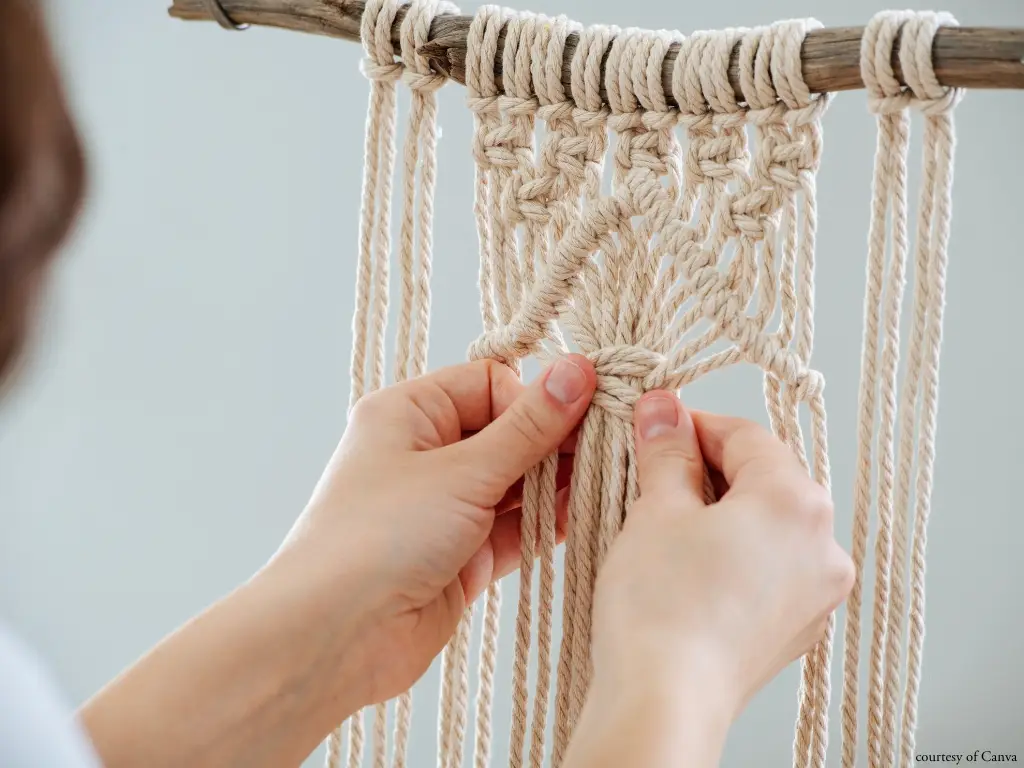 ropes hang from a piece of wood while hands tie knots to create a pattern