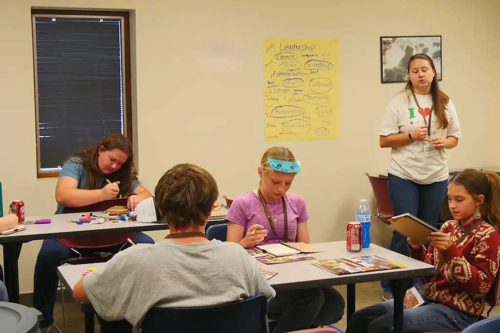 a 4-H leader talks to youth sitting around two tables as they work on a project