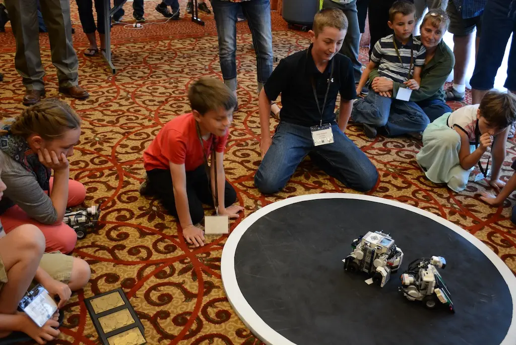 a group of young people sit on the floor and watch two robots competing on a circular mat in front of them