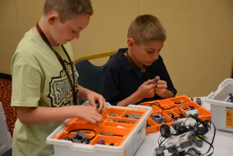 two boys sort through trays of lego parts while building a lego robot that sits in front of them on a table