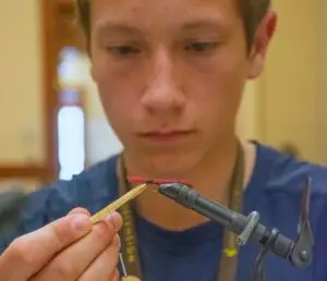 red thread is being added to a hook that is being held in a clamp and tied into a fly. a young man is out of focus in the background