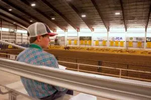 a young person sits in the stands looking out at a dirt arena where someone is riding a horse