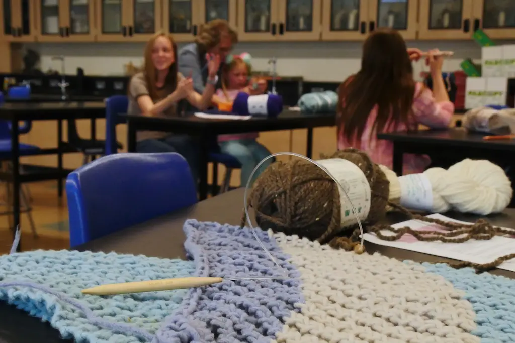 a closeup of a blue and white knitting project with some brown yarn on a table and 3 young people out of focus in the background practicing knitting with the help of an adult