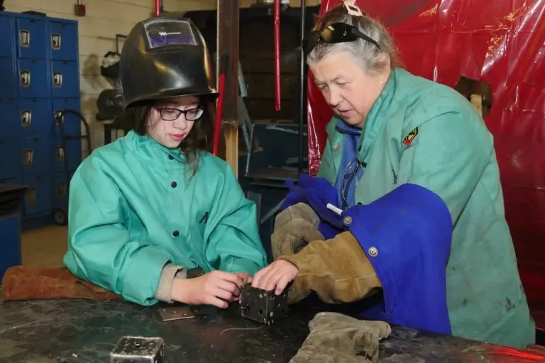 an adult discusses a metal cube that is part of a welding project with a young person. both are wearing welding safety gear.