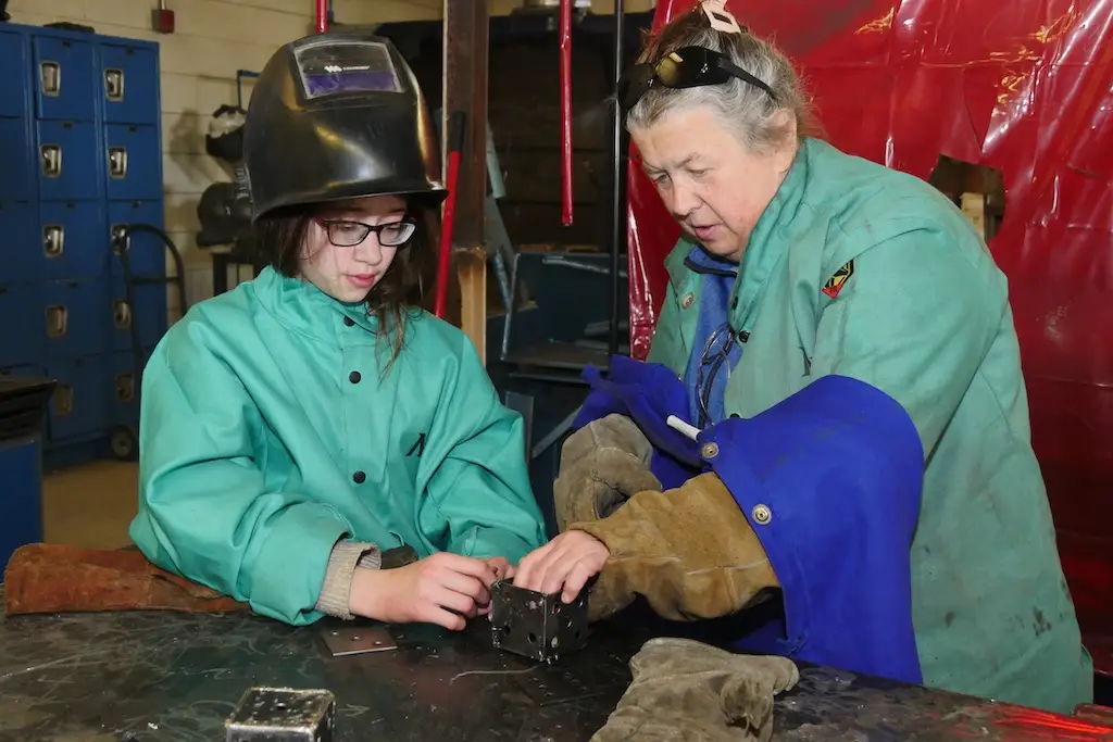 an adult discusses a metal cube that is part of a welding project with a young person. both are wearing welding safety gear.