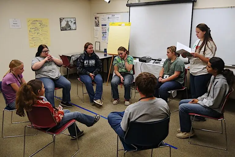 a group of young people sit in a circle while an adult stands and reads from a notebook