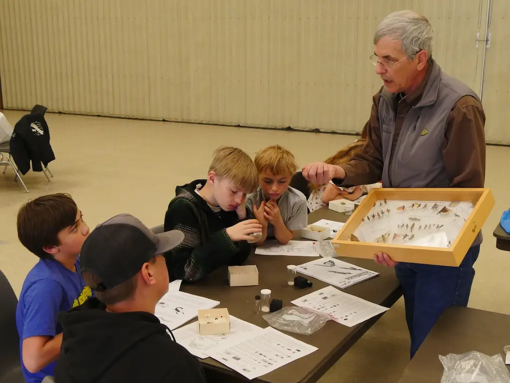 a man holds a case of mounted insects while a group of young people sit at a table in front of him listening