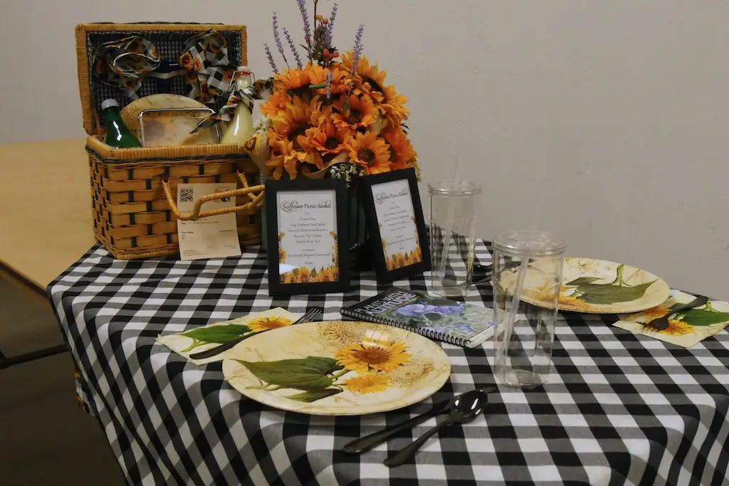 a table setting at the fair that includes plates with sunflowers, a wooden picknick basket, a flower setting of sunflowers, and a checkered black and white table cloth