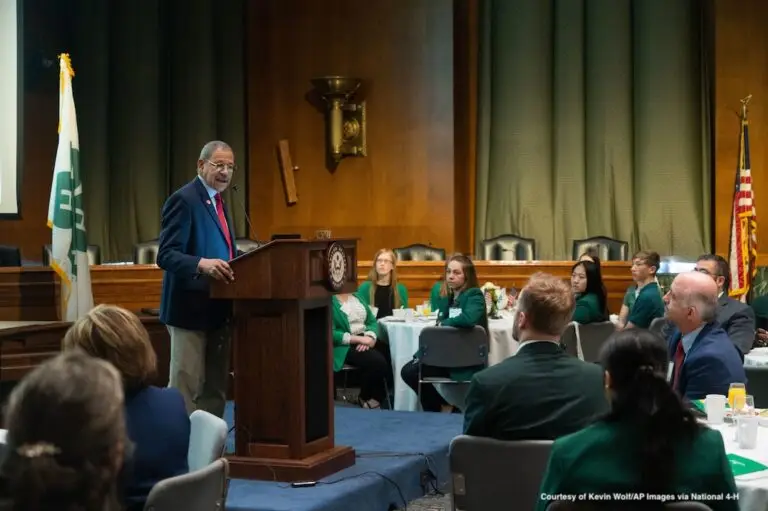 a man stands a a lectern speaking to a group of youth seated at tables