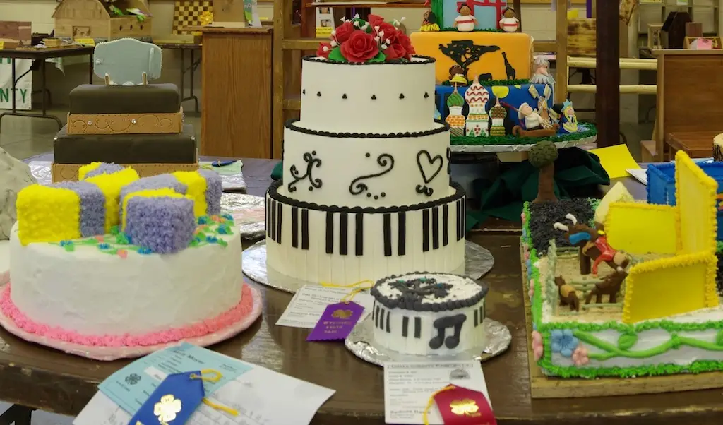 a table displaying cakes at fair including a cake with a butterfly on it, a a cake with piano keys and roses on top and a smaller cake with music notes, and a cake with a rodeo scene