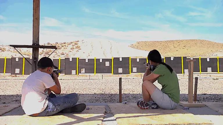 two young people sit on the ground at a rifle range holding rifles to their shoulders ready to shoot. a back stop with targets on it is visible in the background