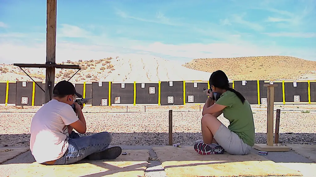 two young people sit on the ground at a rifle range holding rifles to their shoulders ready to shoot. a back stop with targets on it is visible in the background