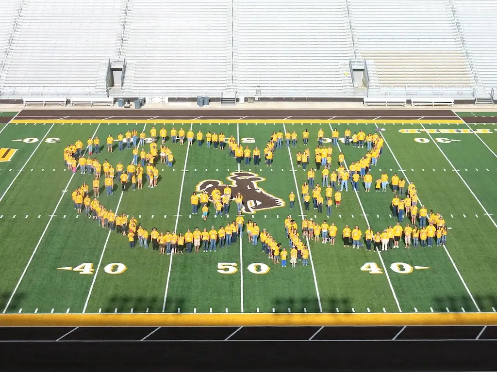 a group of people wearing gold shirts stand on a football field in the shape of a 4-H clover