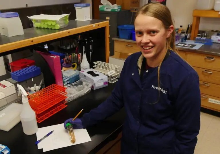 a young woman in a lab coat stands at a lab bench holding a vial of yellow liquid