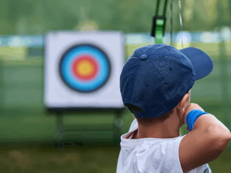 a young person draws a bow and aims at a target out of focus in the background