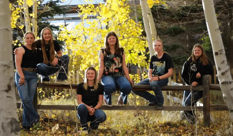 a group of young people sit on a wooden fence posing in front of fall trees