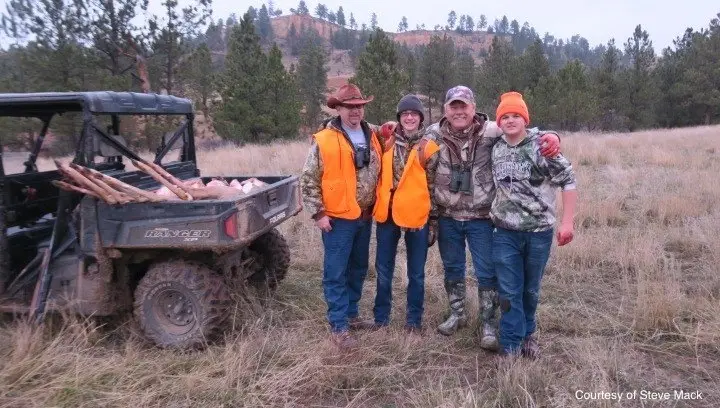 a group of youth and adults in hunter orange pose next to deer in a side by side