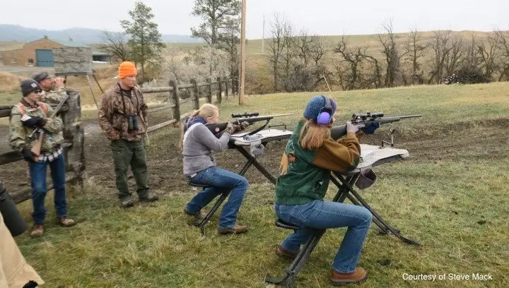 two young women sit at shooting benches siting in their rifles while other youth lean on a fence nearby and watch