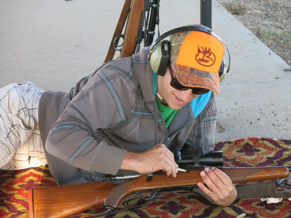 A young person lies prone loading a shell in a rifle