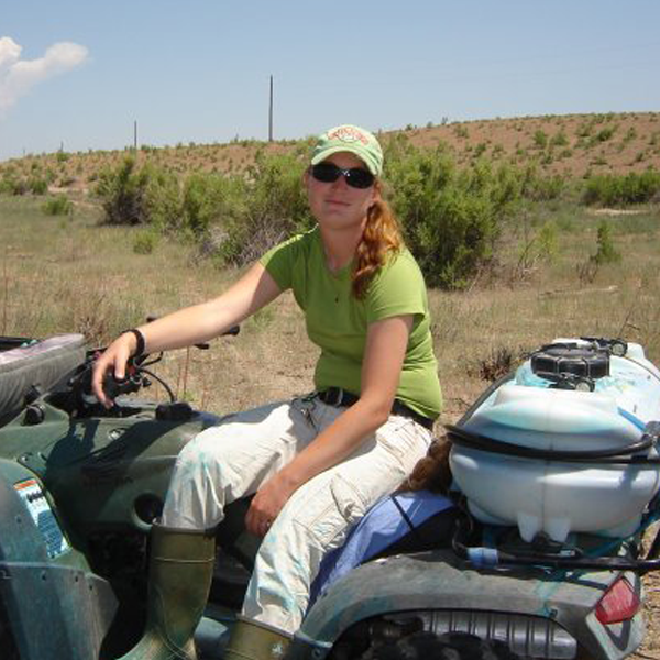 amie Allen wearing a green shirt and beige khakis, riding a 4-wheeler with a pesticide tank on the back